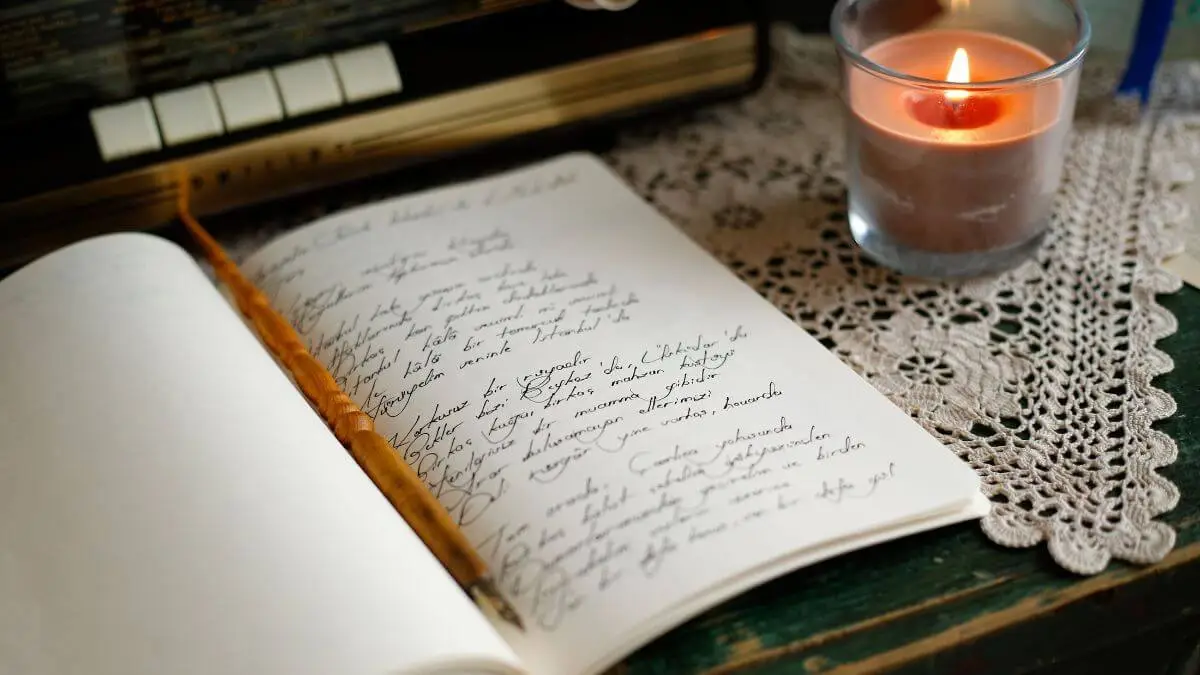 Caderno aberto com anotações manuscritas, pena e vela acesa sobre uma mesa com toalha de renda.