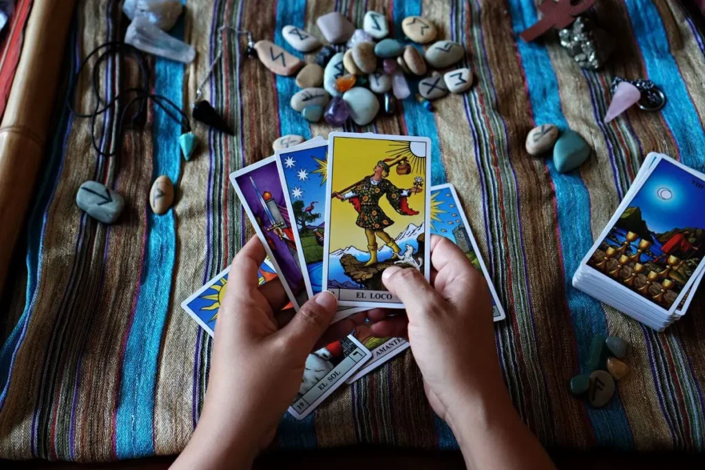Mãos segurando cartas de tarot sobre uma mesa com cristais, pedras, runas e pêndulos.