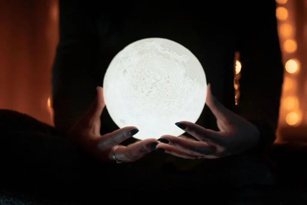 Mãos segurando uma luminária esférica em formato de lua cheia, iluminada em um ambiente escuro e aconchegante.