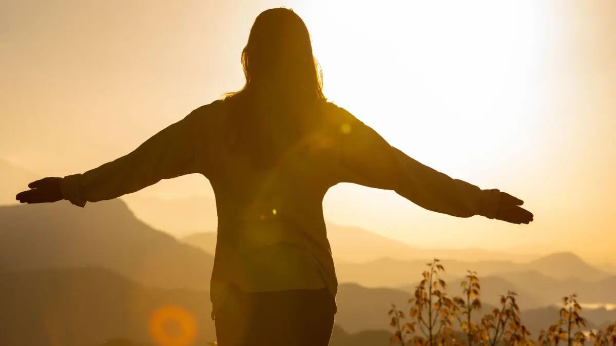 Mulher de braços abertos diante das montanhas, iluminada pela luz dourada do pôr do sol.