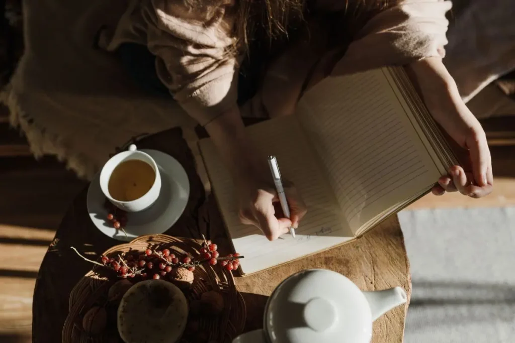Mulher escrevendo em um diário aberto sobre uma mesa de madeira, ao lado de uma xícara de chá e um bule branco.