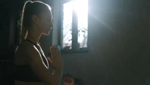 Mulher meditando com os olhos fechados e as mãos unidas diante de uma janela iluminada pelo sol.