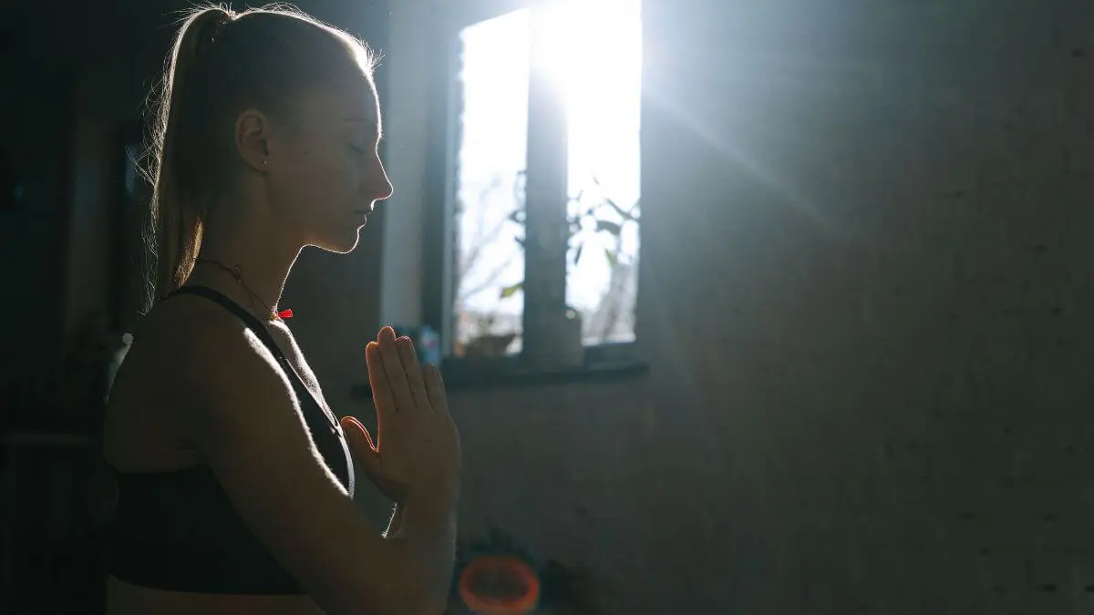 Mulher meditando com os olhos fechados e as mãos unidas diante de uma janela iluminada pelo sol.