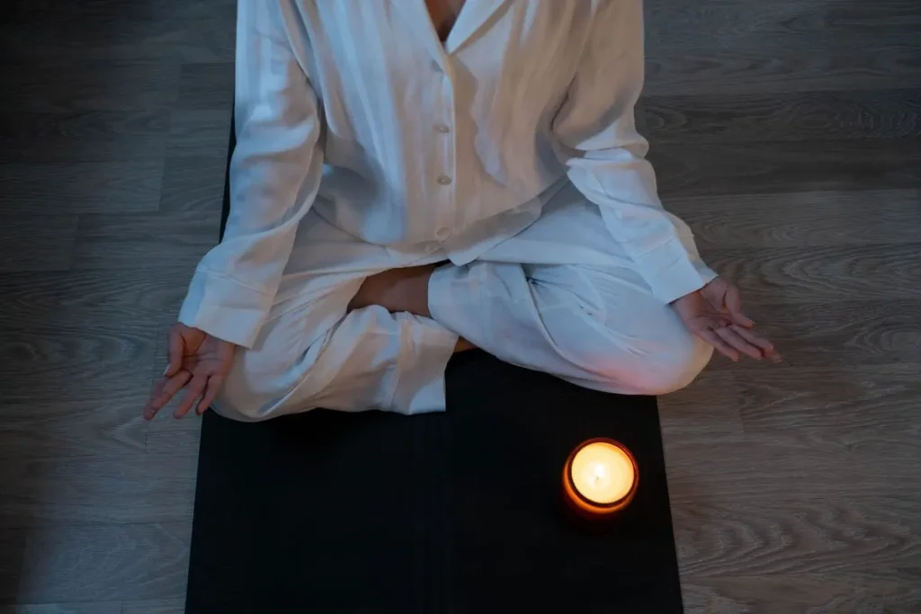 Mulher sentada em posição de meditação sobre um tapete, com uma vela acesa à sua frente.