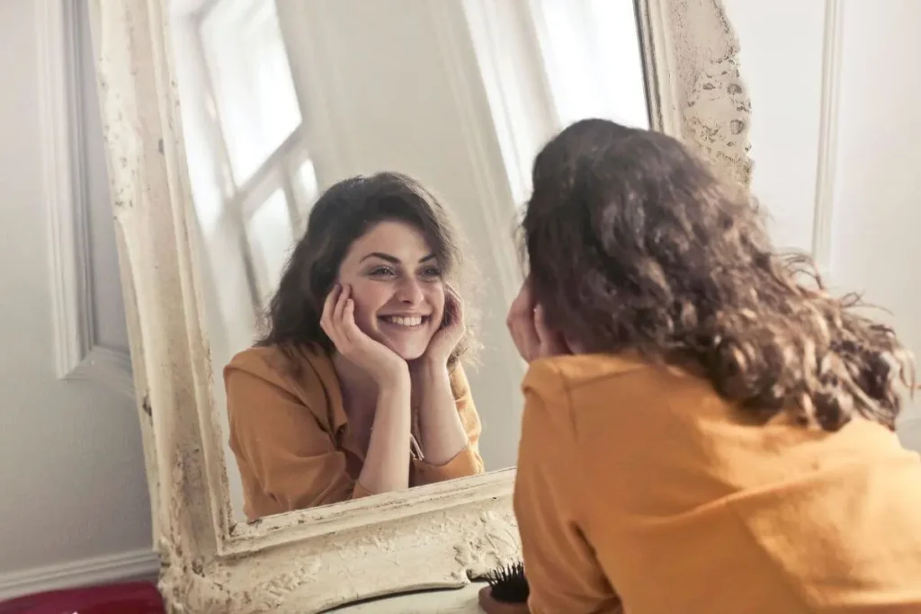Mulher sorrindo para o próprio reflexo no espelho, com as mãos apoiadas no rosto em um ambiente iluminado.