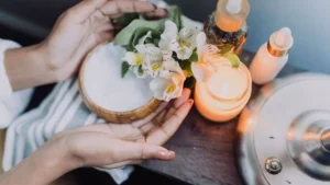 Mãos segurando flores ao lado de uma vela acesa, creme corporal e frascos de cosméticos em um ambiente de autocuidado.