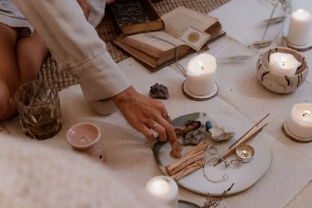 Pessoa organizando cristais, incensos e ervas em um altar com velas acesas e livros antigos.