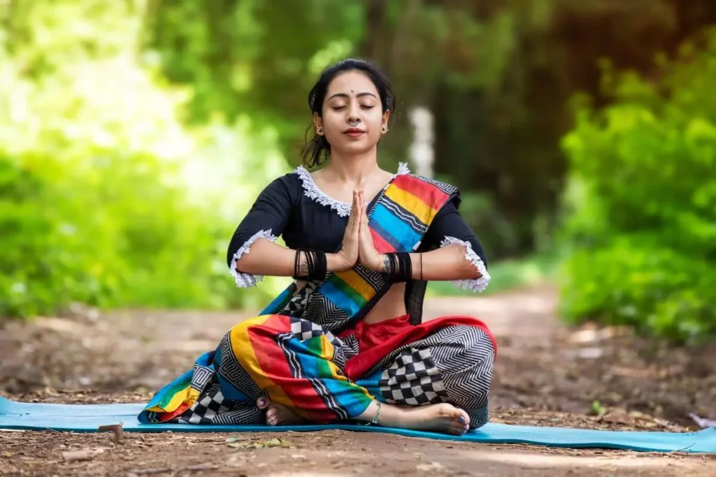 Mulher meditando ao ar livre, sentada em posição de lótus e com as mãos em gesto de oração.