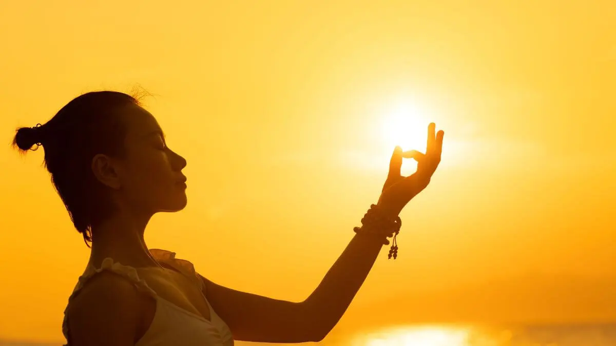 Silhueta de mulher meditando ao pôr do sol com a mão direcionada para a luz do sol.